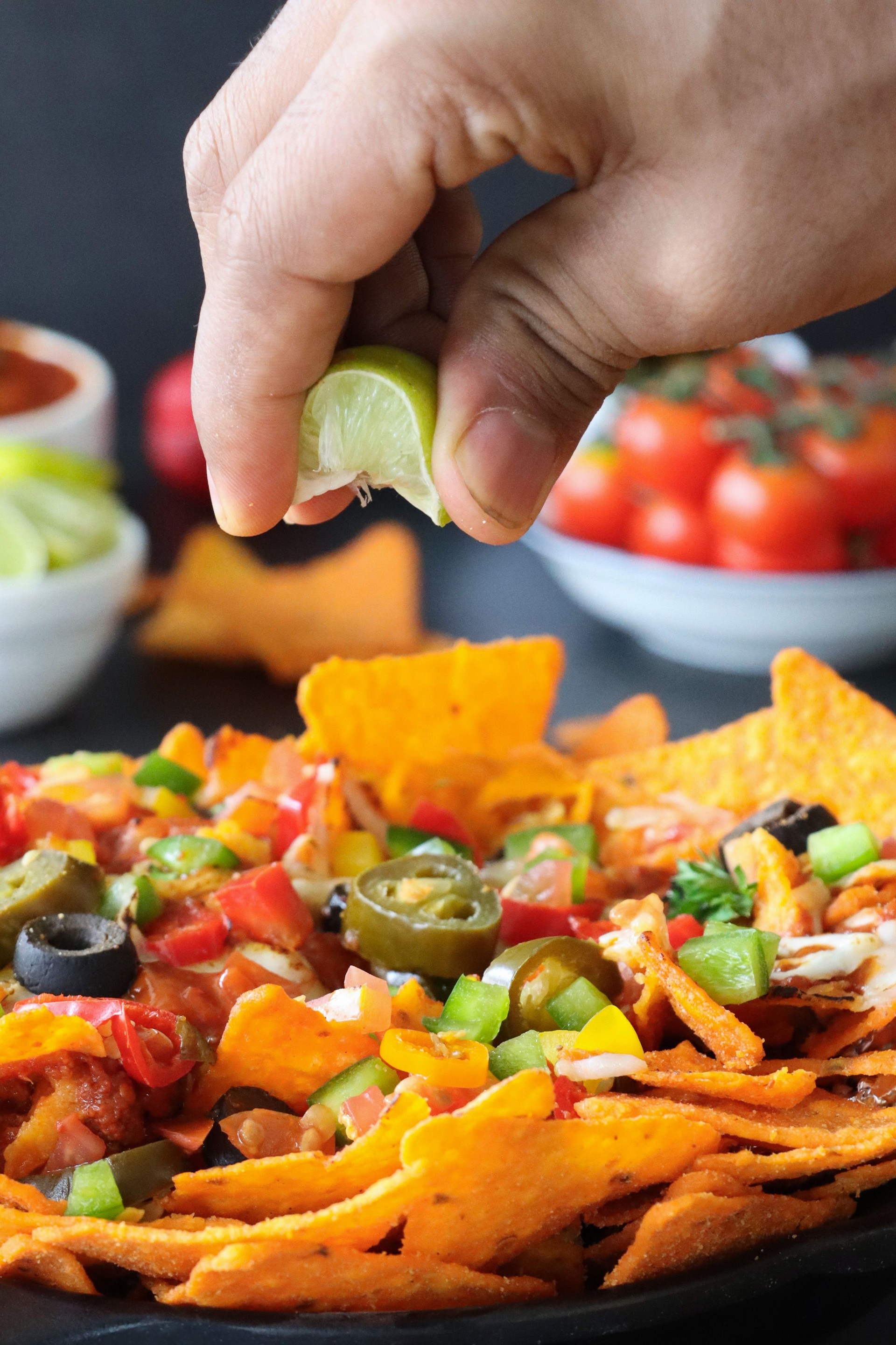 Full frame image of hand squeezing lime wedge over homemade, loaded nachos covered in melted mozzarella cheese, cherry tomatoes, black olives, Jalapeno peppers, elevated view, focus on foreground, served with sauces and dips